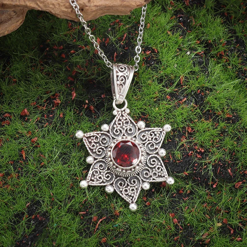 Silver star-shaped pendant with a red gemstone on a chain, set against a natural background.
