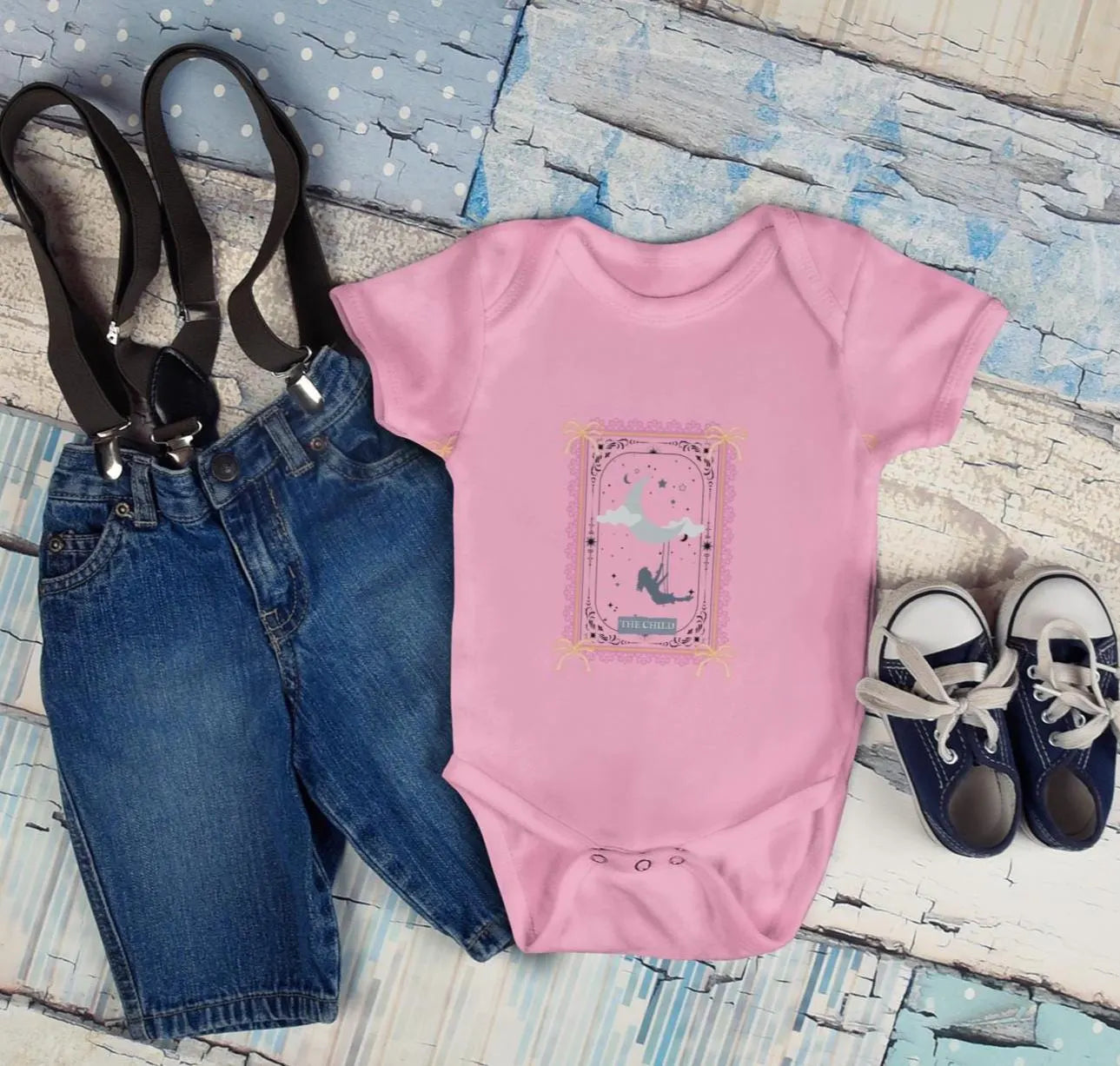 Pink baby onesie with a design, blue jeans, suspenders, and blue shoes on a textured surface.