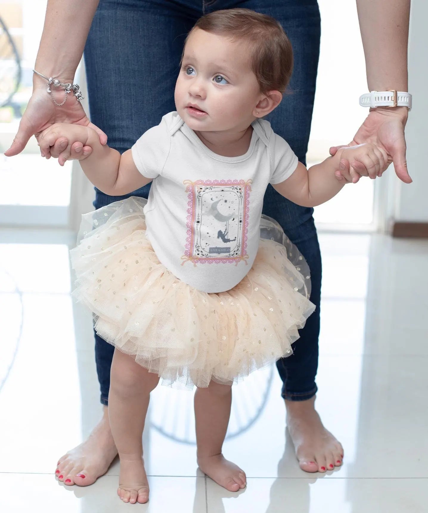 Baby in a tutu standing with adults holding hands indoors
