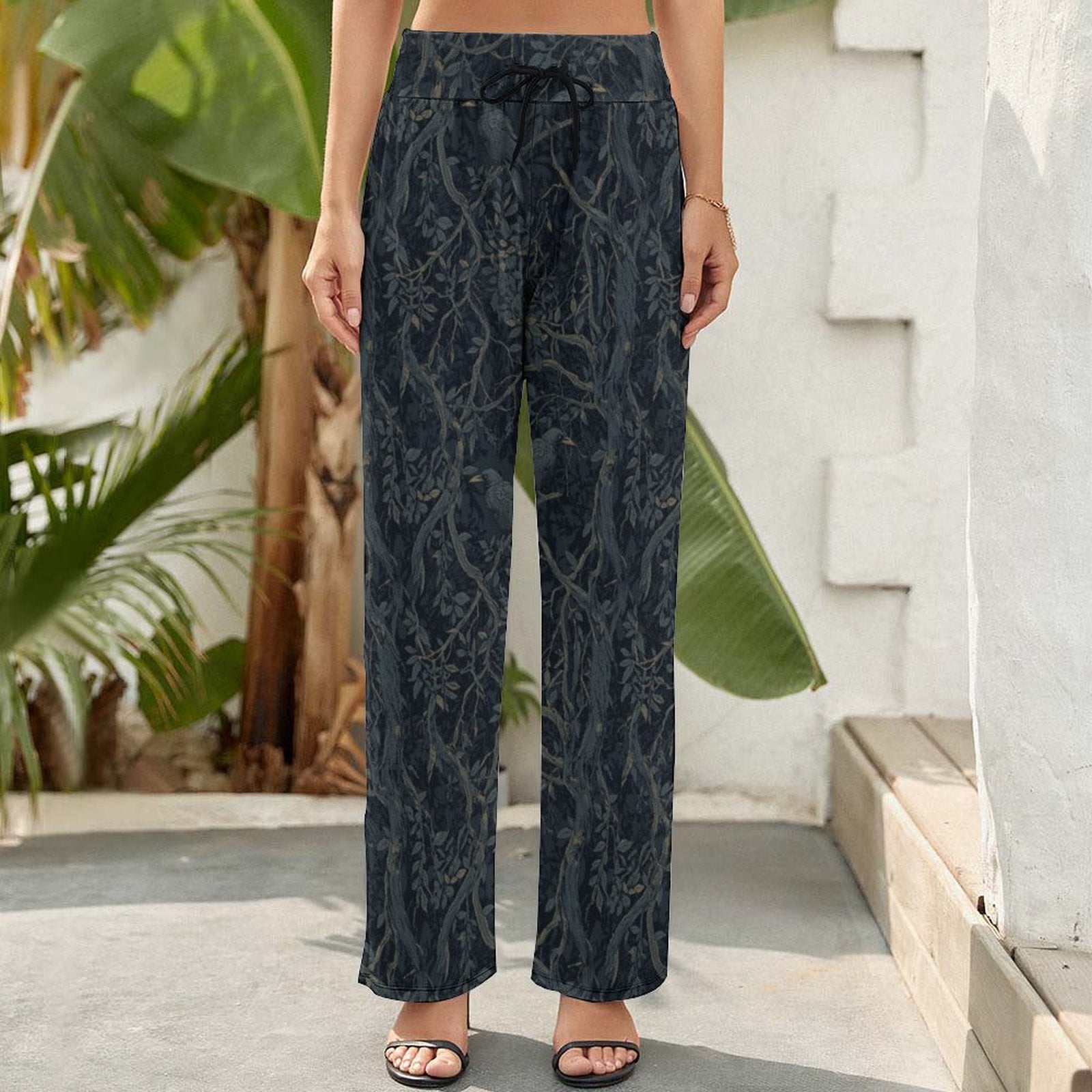 Person wearing dark patterned pants standing outdoors with plants and a stone wall in the background