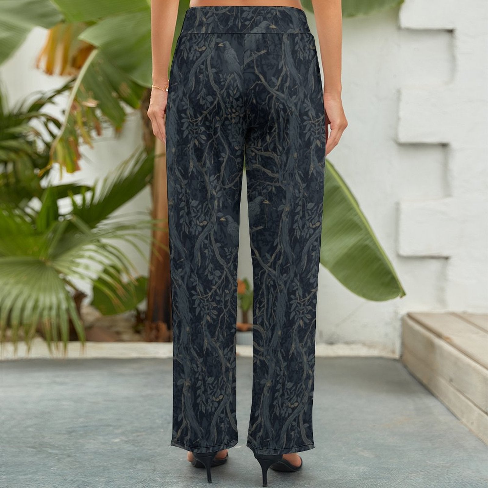 Person wearing dark patterned pants standing outdoors with plants and a wall in the background