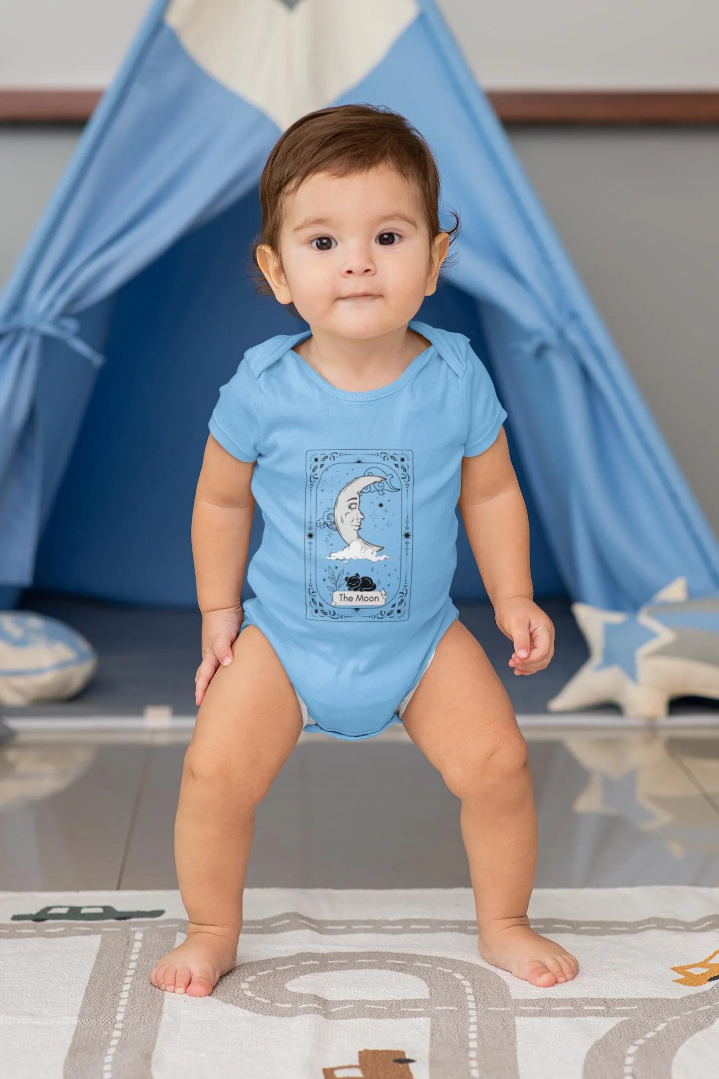Baby in a blue onesie standing in front of a blue teepee.
