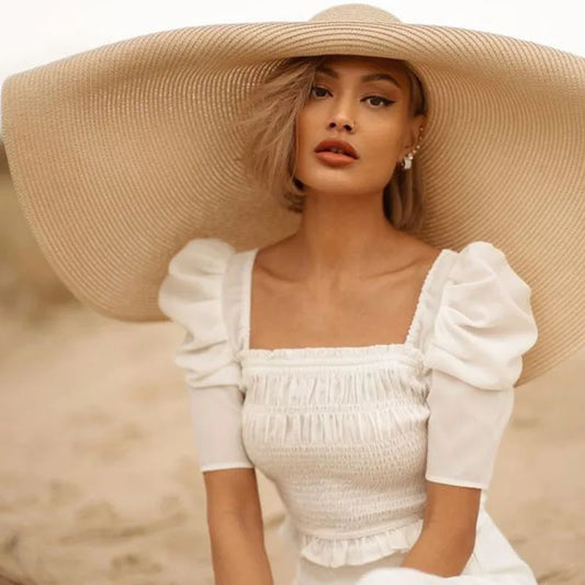 Woman wearing a white dress and large beige hat against a neutral background