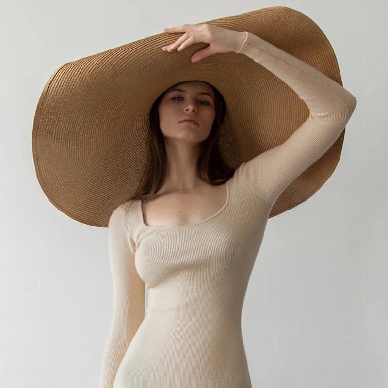 Woman wearing a beige long-sleeve top and large brown hat against a plain background