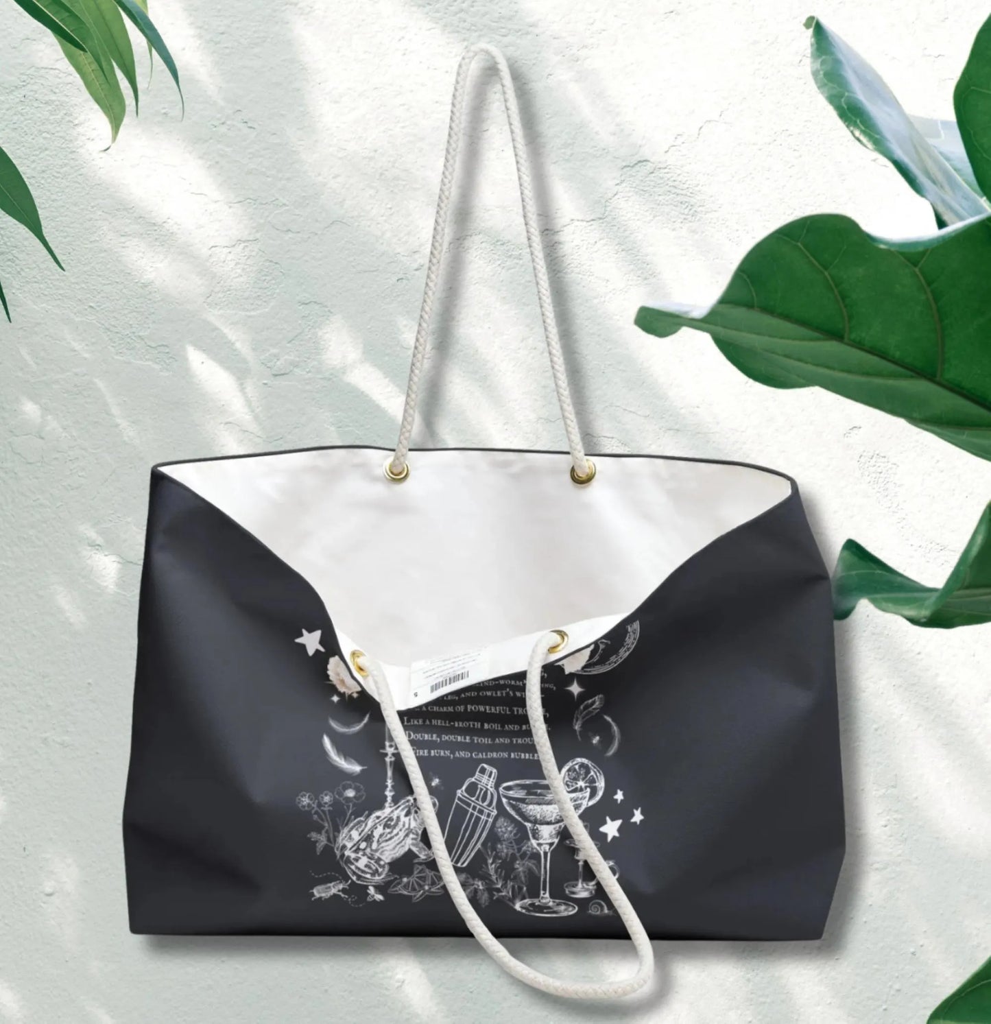 Black tote bag with white handle and graphic design on a light gray background with green leaves.