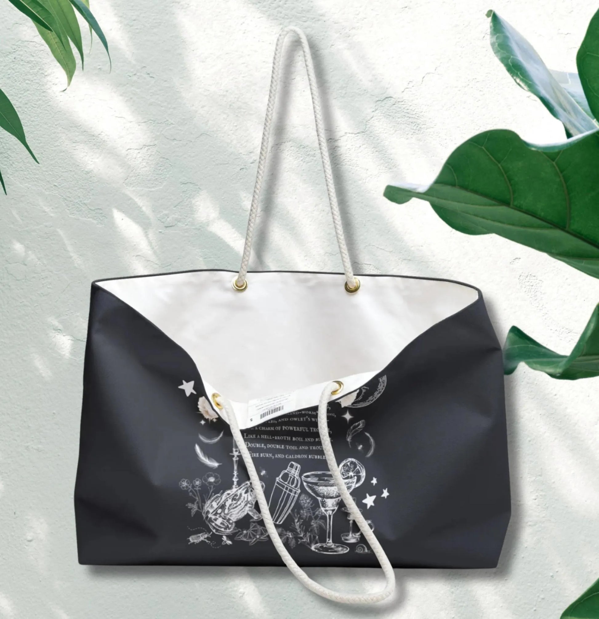 Black tote bag with white handle and graphic design on a light gray background with green leaves.
