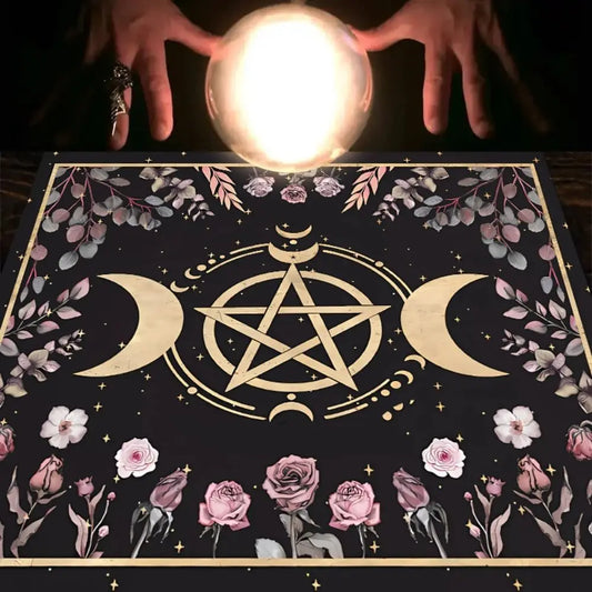 Crystal ball held by hands on a table with a decorative cloth featuring floral and pentagram designs.