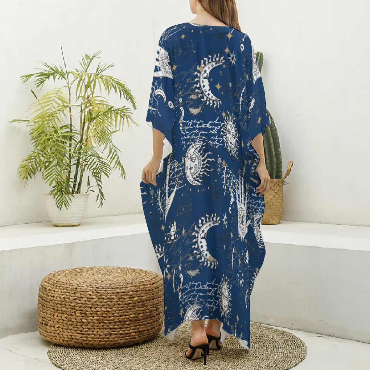 Woman wearing a blue patterned dress standing in a room with a plant and woven ottoman.