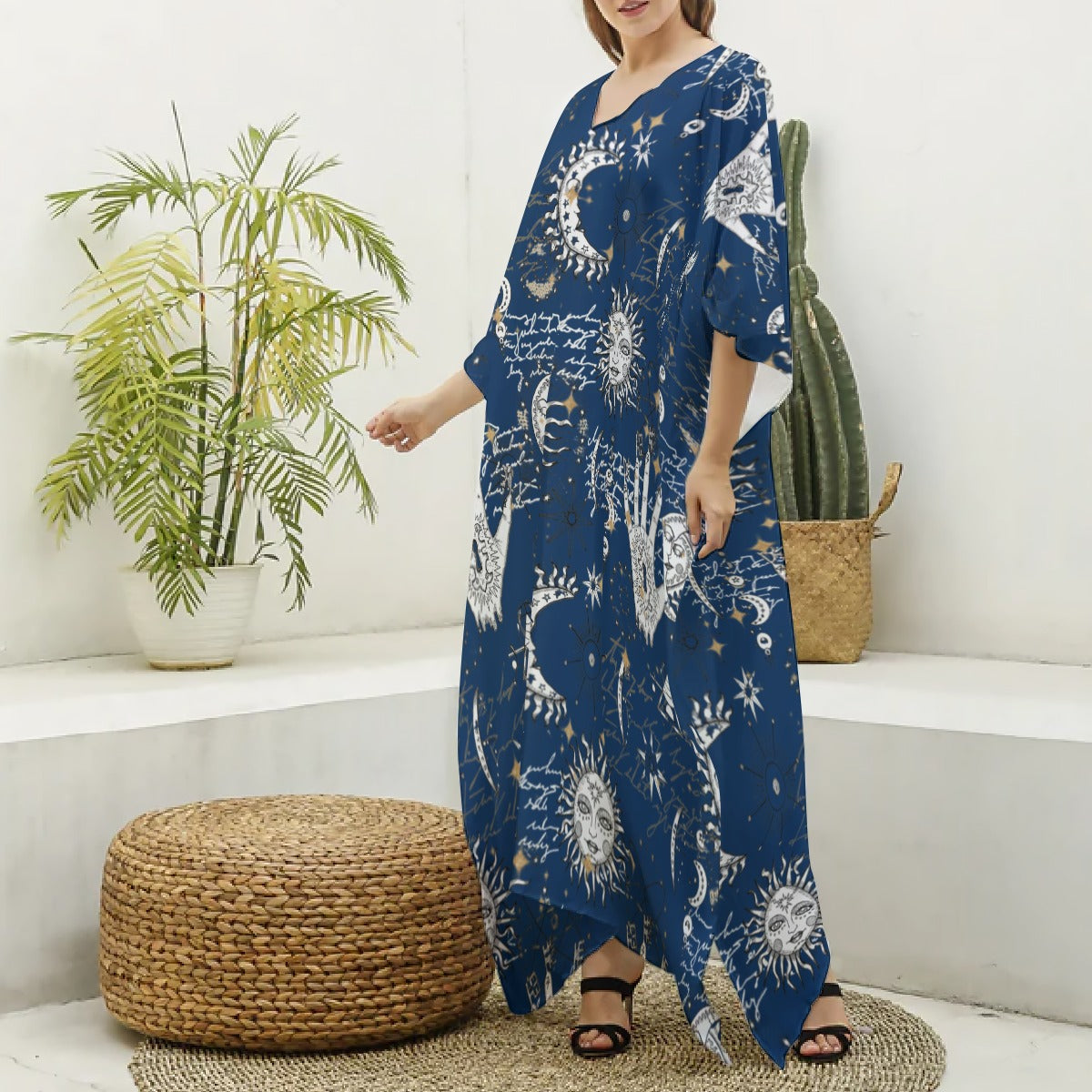 Woman wearing a blue patterned dress standing in a room with plants and a woven ottoman.