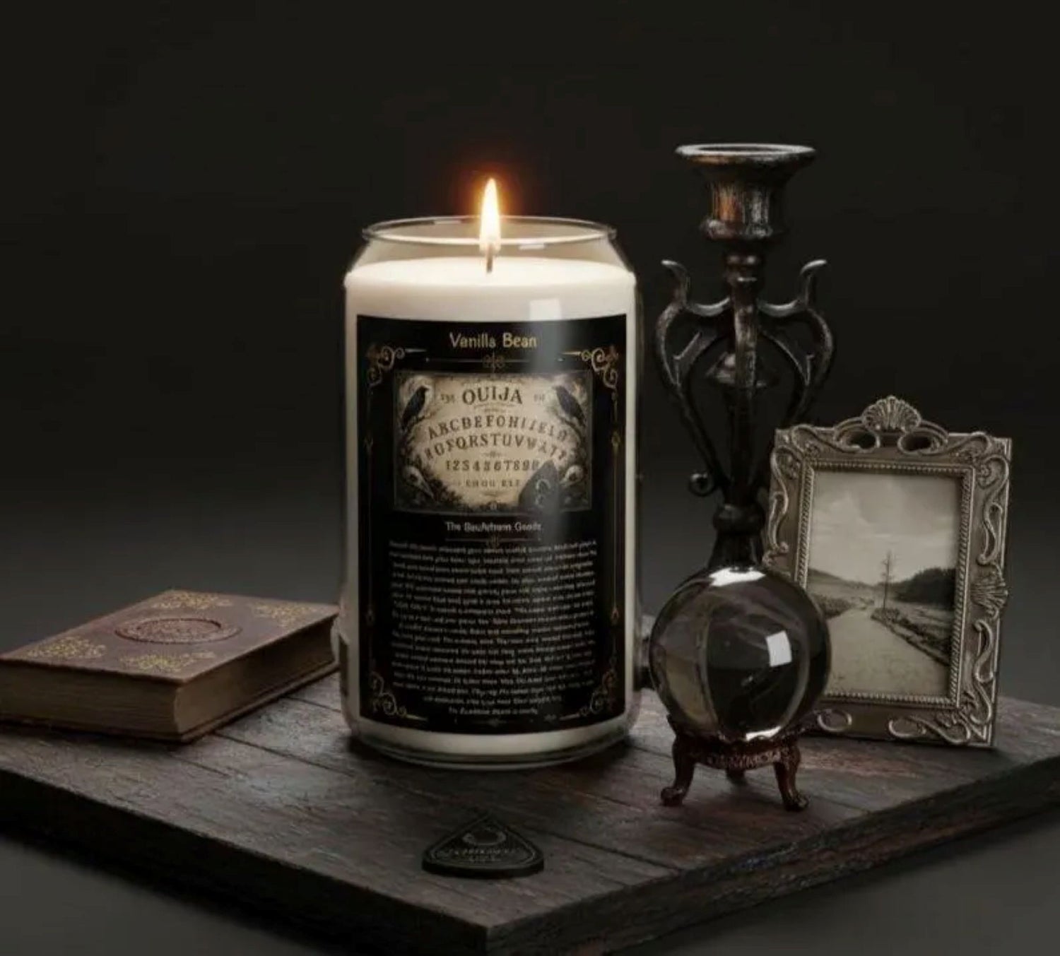 Candle with a label on a dark surface with a book, bottle, and picture frame.