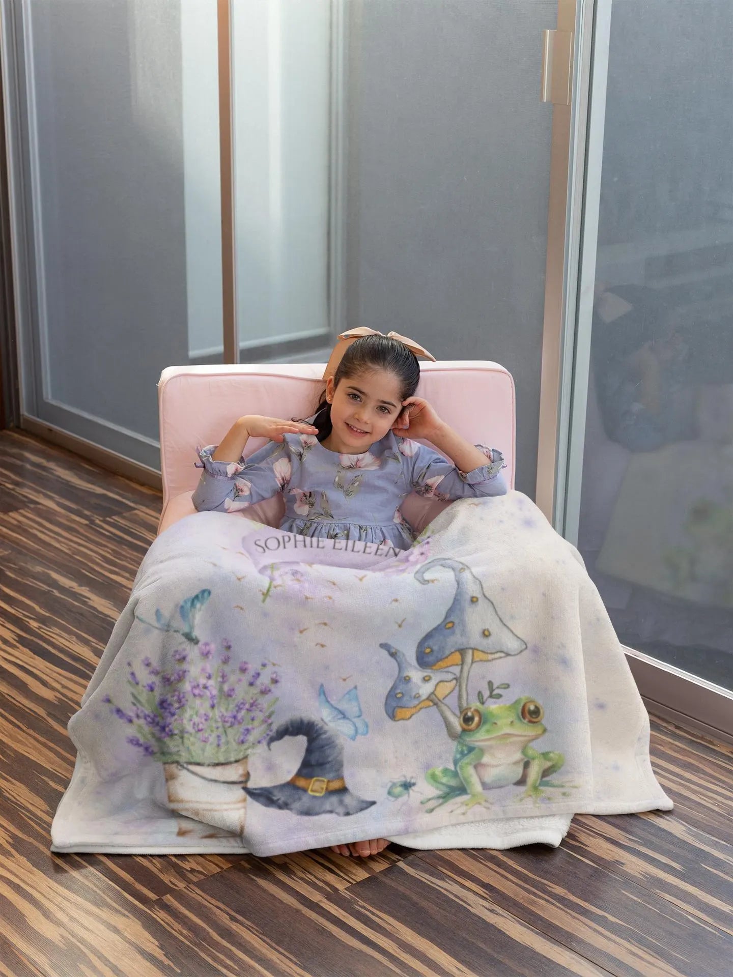 Child sitting on a chair with a blanket featuring cartoon characters, in a room with large windows.