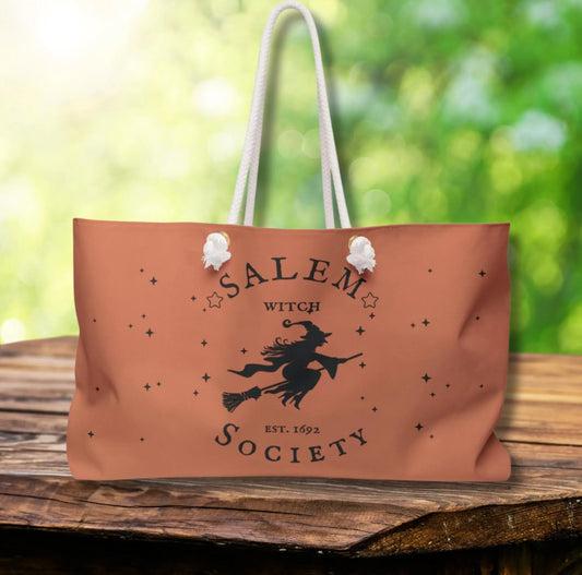 Tan tote bag with 'Salem Witch Society' design on a wooden surface with a blurred green background