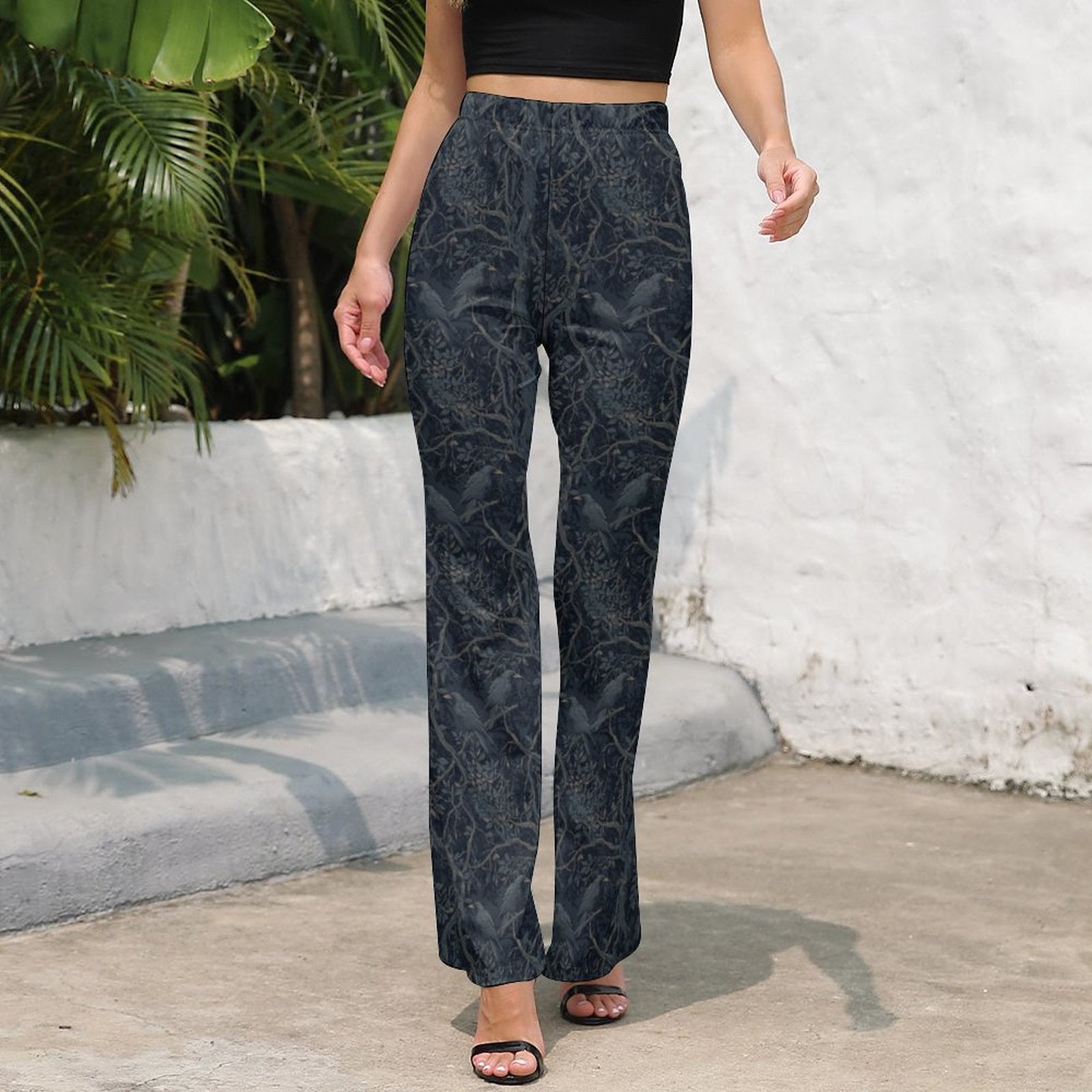 Person wearing dark patterned pants standing outdoors with a white wall and greenery in the background