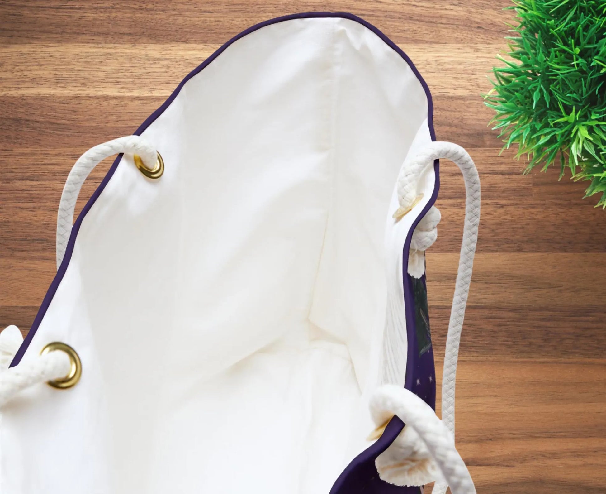 White drawstring bag with gold accents on a wooden surface with a plant in the corner