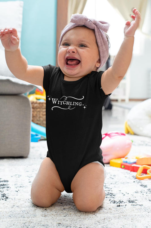 Baby wearing a black onesie with 'Witchling' text, sitting on a carpeted floor with toys around.