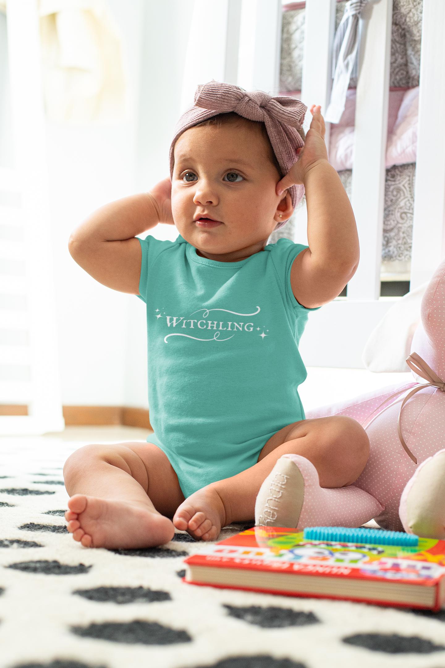Baby wearing a teal onesie with 'Witchling' text, sitting on a patterned rug.