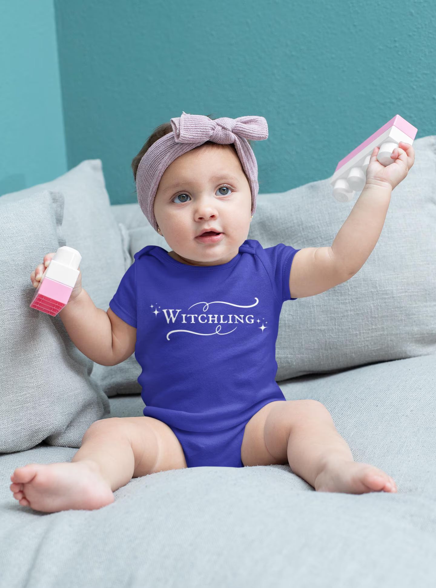 Baby wearing a purple onesie with 'Witchling' text, sitting on a couch.