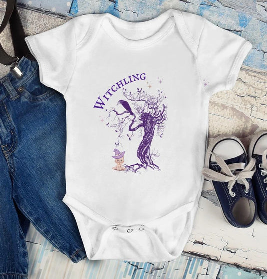 White baby onesie with purple graphic and text 'Witchling' on a textured surface with jeans and shoes.