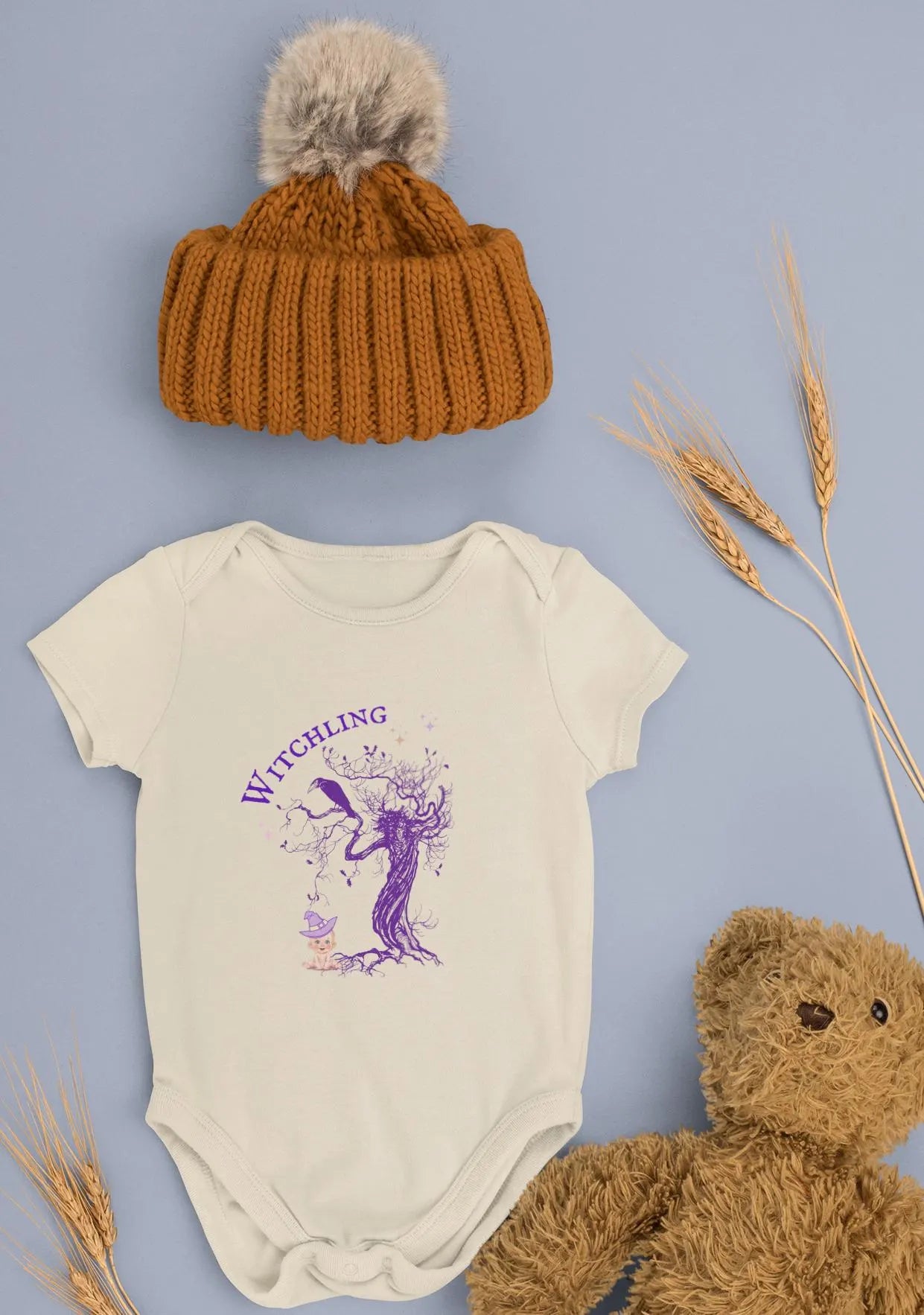 Baby onesie with 'Witchling' design, brown knit hat, and teddy bear on a light blue background.