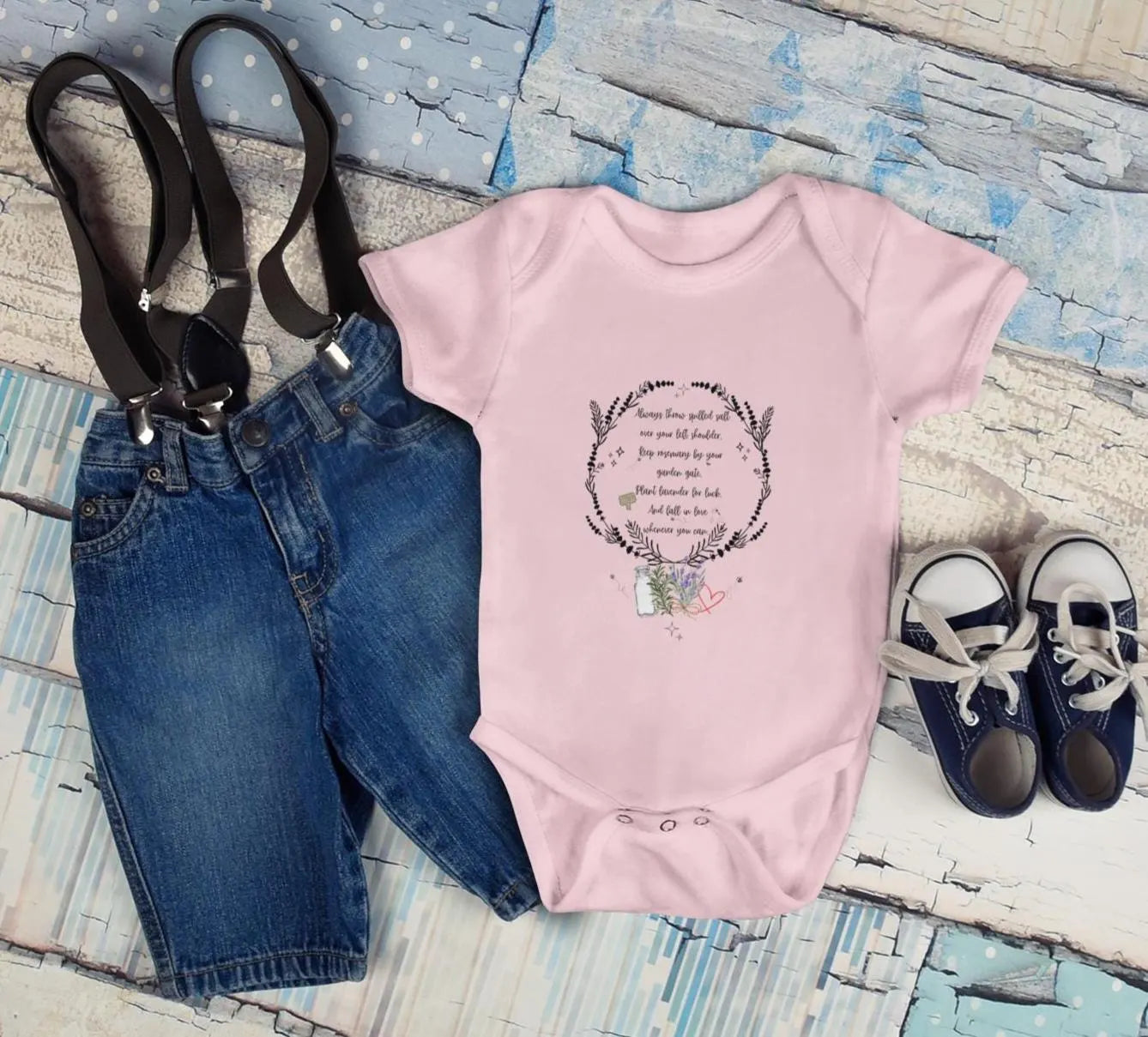 Pink baby onesie with text, blue jeans, and shoes on a textured surface